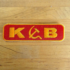 Könnte beinhalten: Ein roter gestickter Aufnäher mit den Buchstaben "K" und "B" in Gelb, mit einem gelben Hammer-und-Sichel-Symbol zwischen den Buchstaben.