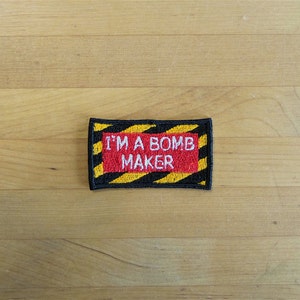 Op de afbeelding: Een zwart-gele gestreepte patch met de tekst "I'M A BOMB MAKER" in witte letters op een rode achtergrond.
