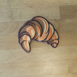 May include: A brown and orange embroidered patch of a croissant. The croissant is a popular French pastry.