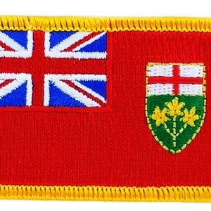 May include: A red embroidered patch with a yellow border. The patch features a Union Jack in the top left corner and a green shield with a white cross and three yellow flowers in the bottom right corner.