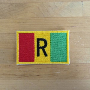 May include: A yellow, red, and green embroidered patch with the letter "R" in black on a yellow background.