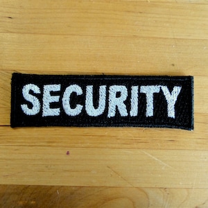 Può includere: Toppa in tessuto nero con testo ricamato in bianco che recita "SECURITY".