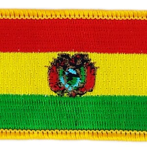 May include: A rectangular embroidered patch with the flag of Bolivia. The flag features three horizontal stripes: red, yellow, and green. The yellow stripe has the Bolivian coat of arms in the center. The patch has a yellow border.