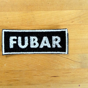 Peut inclure: Écusson rectangulaire noir avec le mot "FUBAR" brodé en blanc. L'écusson a une bordure blanche et est posé sur une surface en bois brun clair. L'écusson est rectangulaire.