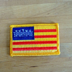 May include: A yellow and red embroidered patch with a blue canton featuring a white design of four castles. The patch is a representation of the Catalan flag.