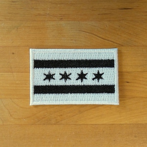 Può includere: Una toppa bianca con una bandiera di Chicago ricamata in nero. La bandiera ha sei stelle bianche su sfondo nero.