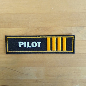 May include: Black embroidered patch with white text "PILOT" and four yellow stripes.