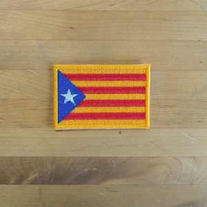 Puede incluir: Un parche bordado amarillo y rojo con un triángulo azul y una estrella blanca en la esquina. El parche está sobre una superficie de madera.