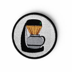 May include: Embroidered patch featuring a coffee maker design. The patch is circular with a white background and a black border. The coffee maker is depicted in black, gray, and gold, with a green accent. A fun accessory for coffee lovers.
