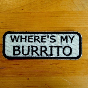 May include: A white embroidered patch with black stitching that reads "Where's My Burrito?" The patch is rectangular and has a black border.