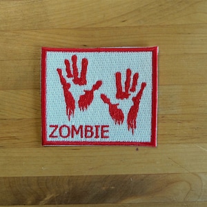 Op de afbeelding: Rode geborduurde patch met twee bloederige handafdrukken en het woord "ZOMBIE" in wit.