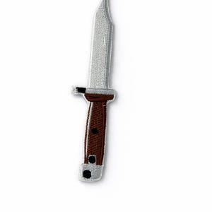 Op de afbeelding: Geborduurde patch van een bajonet met een zilveren lemmet en een bruin handvat. Het handvat heeft twee zwarte stippen en een zwart accent bij het lemmet. De patch staat op een witte achtergrond.