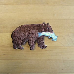 Könnte beinhalten: Gestickter Aufnäher eines braunen Bären, der einen Fisch im Maul trägt. Der Bär ist braun und der Fisch ist weiß mit rosa und blauen Akzenten.