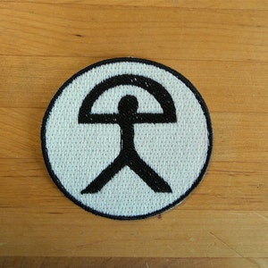 Op de afbeelding: Een witte ronde patch met een zwart symbool van een gestileerde menselijke figuur met een gebogen lijn boven zijn hoofd.