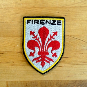 May include: A white and yellow embroidered patch with a red fleur-de-lis and the text "FIRENZE" in black.