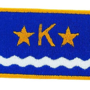 May include: Embroidered patch with a blue background, featuring two gold stars and a gold letter "K". A white wavy line is at the bottom. The patch has a gold border.