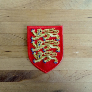 May include: A red embroidered patch with three gold lions rampant, a traditional symbol of England.