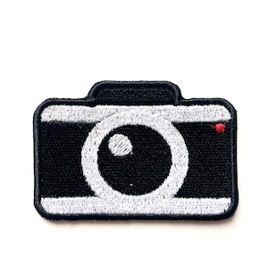 Peut inclure: Un patch thermocollant brodé noir et blanc représentant un appareil photo. L'appareil photo a un objectif blanc et un point rouge dans le coin supérieur droit.