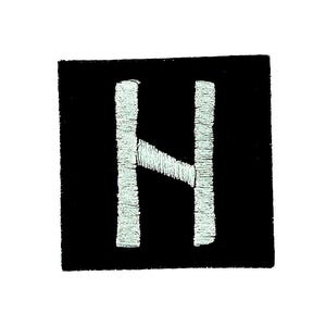 Puede incluir: Un parche cuadrado negro con la letra "H" bordada en blanco. La letra está cosida con un hilo texturizado, ligeramente blanquecino. Este parche es probablemente para ropa o accesorios.