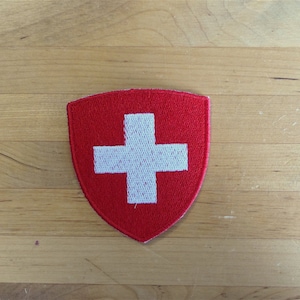 Puede incluir: Un parche de fieltro rojo en forma de escudo con una cruz blanca en el centro. El parche es una representación de la bandera suiza.