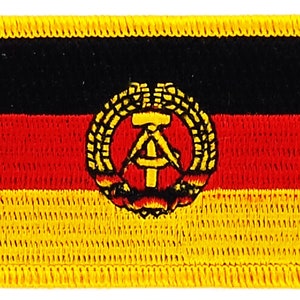 Puede incluir: Un parche bordado con la bandera de la República Democrática Alemana (RDA). El parche es negro, rojo y amarillo con un martillo y una brújula dentro de una corona de trigo.