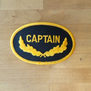 Könnte beinhalten: Schwarzer ovaler Aufnäher mit gelber gestickter Aufschrift "CAPTAIN" und zwei gelben gestickten Lorbeerkränzen.