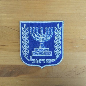 May include: A blue and white embroidered patch depicting the Israeli emblem. The design includes a menorah, olive branches, and Hebrew text. The patch is shield-shaped and set against a wooden background.