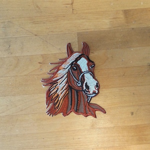 May include: Embroidered patch of a brown and white horse's head with a bridle. The horse has a white star on its forehead and a white stripe down its nose.