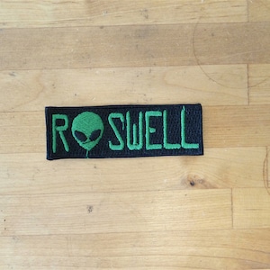 May include: A black and green embroidered patch with the word "ROSWELL" and a green alien head.