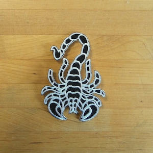 Puede incluir: Parche bordado de escorpión en blanco y negro. El escorpión está mirando hacia la derecha con la cola enroscada. El parche está sobre una superficie de madera.