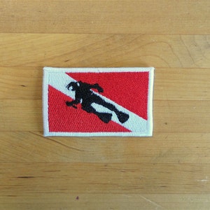 May include: A white and red scuba diving flag with a black silhouette of a diver. The flag is rectangular and has a white border.