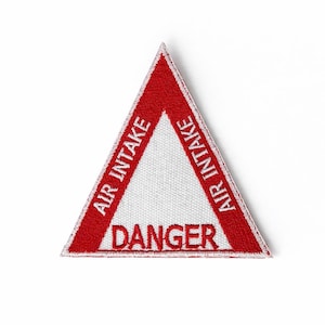 Puede incluir: Un parche triangular rojo y blanco con las palabras "AIR INTAKE" en los lados y "DANGER" en el centro. El parche tiene un fondo blanco y un borde rojo.