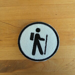 Peut inclure: Patch rond blanc avec un contour noir. Le patch présente une silhouette noire d'une personne avec un sac à dos et un bâton de marche.