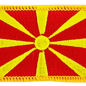 May include: Embroidered patch featuring the flag of North Macedonia. The design includes a red background with a yellow sunburst radiating from the center. The patch has a yellow border.