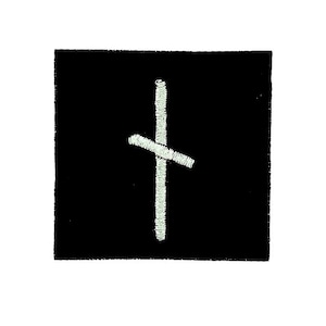 May include: A square black patch with a white embroidered symbol. The symbol is a vertical line intersected by a diagonal line, resembling a stylized cross. The patch has a textured appearance.
