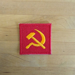 Könnte beinhalten: Ein roter Stoff-Patch mit einem gestickten Hammer und Sichel in Gelb.