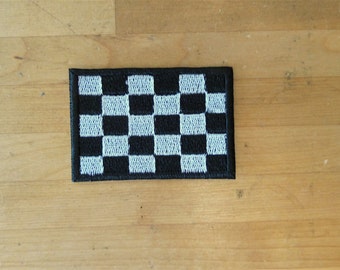 May include: A black and white checkered flag patch. The patch is embroidered with white stitching on a black fabric background.