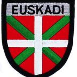 May include: Embroidered patch featuring the Basque flag, a red field with a white cross and green diagonal stripes. The word "EUSKADI" is embroidered in white letters across the top. The patch has a black border.