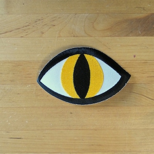 Könnte beinhalten: Ein schwarzer und gelber gestickter Augen-Patch mit weißem Zentrum. Der Patch ist augenförmig und hat einen schwarzen Umriss.