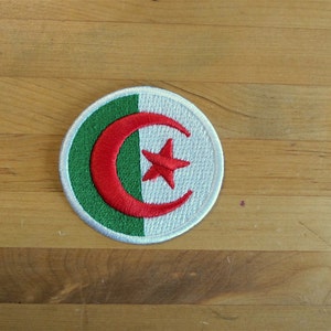 May include: A round embroidered patch featuring the flag of Algeria. The patch depicts a white crescent moon and a red five-pointed star on a green background.