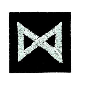 Può includere: Una toppa quadrata nera con un disegno ricamato bianco. Il disegno è una forma simmetrica, simile a una 'X' o a una freccia a doppia punta. Il ricamo ha un aspetto strutturato.