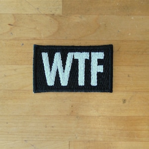 Può includere: Una toppa nera con lettere ricamate bianche che compongono la scritta "WTF"