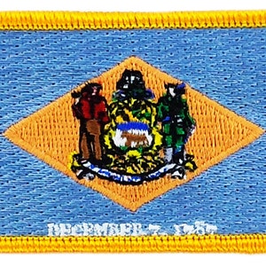 May include: A blue and yellow embroidered patch featuring the state seal of Delaware. The seal depicts a farmer holding a plow, a soldier with a musket, and a ship. The text "DECEMBER 7, 1787" is visible at the bottom of the patch.