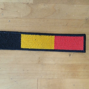 May include: A black, yellow, and red embroidered patch with a black border. The patch is rectangular and has the colors of the Belgian flag.