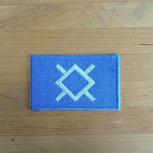 Puede incluir: Un parche de tela azul con un símbolo bordado en blanco. El símbolo es una forma de diamante con cuatro líneas que se extienden desde las esquinas.
