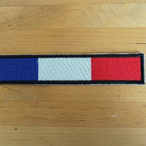 Puede incluir: Un parche bordado azul, blanco y rojo con un borde negro. El parche tiene forma de rectángulo y mide aproximadamente 5 cm de largo y 2,5 cm de ancho.