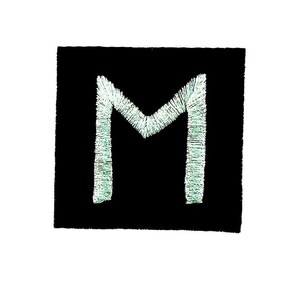 Può includere: Una toppa quadrata nera con la lettera "M" verde chiaro ricamata al centro. La lettera è realizzata con filo strutturato, creando un effetto in rilievo sullo sfondo scuro. La toppa ha una forma leggermente irregolare.