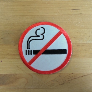Puede incluir: Un parche redondo blanco con borde rojo que muestra un símbolo de prohibido fumar. El símbolo incluye un cigarrillo con humo y una línea diagonal roja. El parche está sobre una superficie de madera.