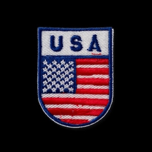 Könnte beinhalten: Gestickter Aufnäher mit dem Design der amerikanischen Flagge und dem Text "USA" in weißen Buchstaben. Der Aufnäher hat ein rot-weiß-blaues Farbschema und ist vor einem schwarzen Hintergrund platziert.