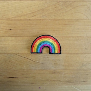 Può includere: Una toppa ricamata a forma di arcobaleno. La toppa è bordata con filo nero e presenta uno schema di colori rosso, arancione, giallo, verde, blu, indaco e viola.